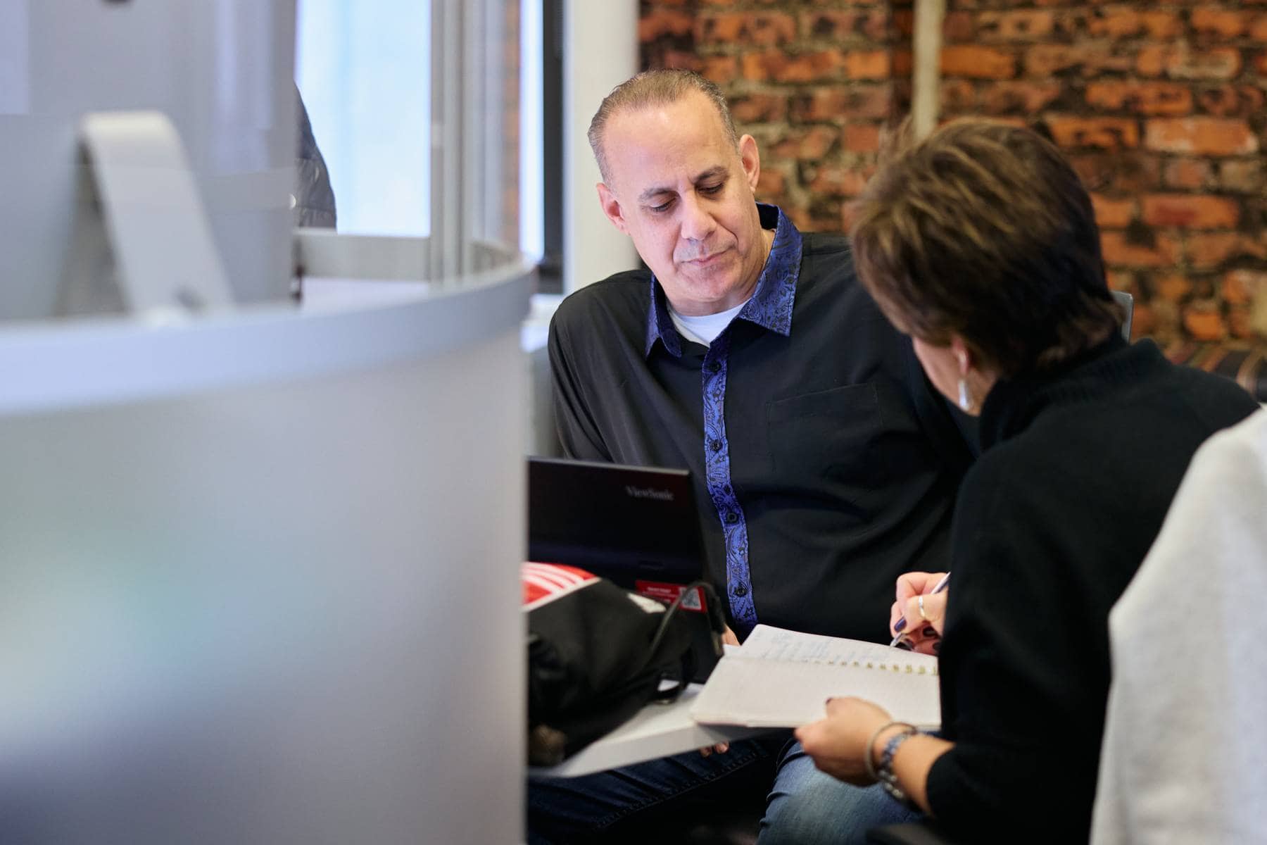 A man and woman engaged in discussion at a table, focusing on a notebook. Brick wall backdrop suggests a cozy, casual office environment.